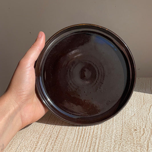 Hand holding a dark ceramic plate on a textured surface with a neutral background