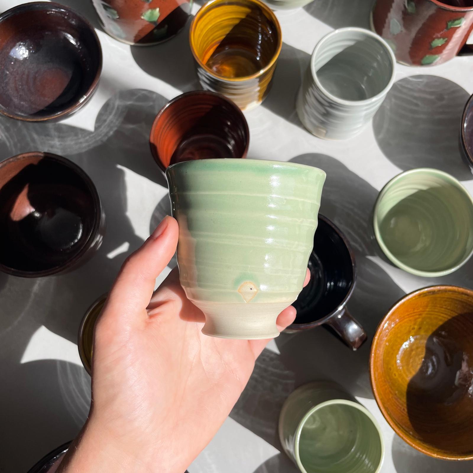 Hand holding a green ceramic cup amidst various other ceramic items on a light surface.