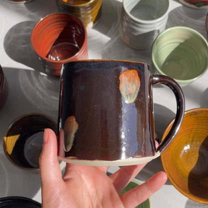Hand holding a brown ceramic mug with other mugs in the background on a light surface.