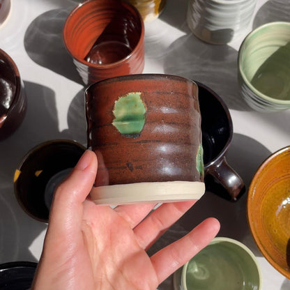 Hand holding a ceramic cup with a green design against a background of various ceramic items.